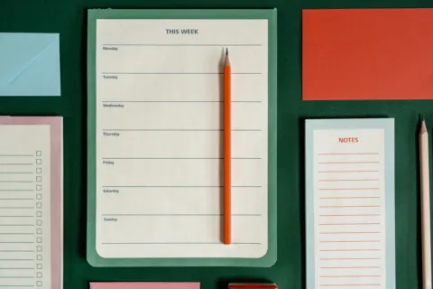 A blank weekly planner pad headed "This week" with an orange pencil on it, laid on a dark green surface among notepads.