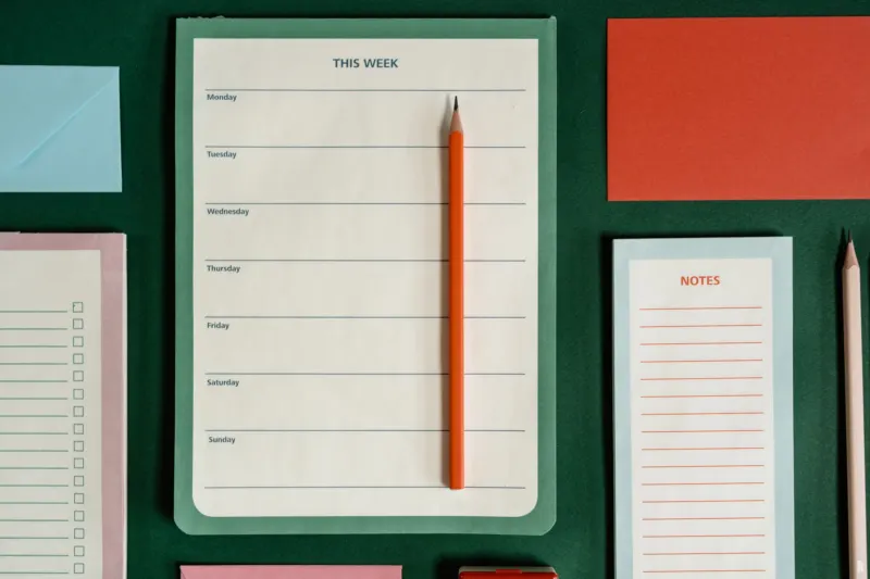 A blank weekly planner pad headed "This week" with an orange pencil on it, laid on a dark green surface among notepads.