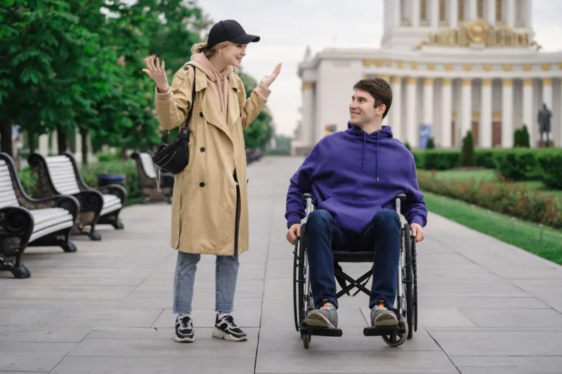 A man propelling his wheelchair and a woman walking beside him chat and smile as they move along a wide park path.