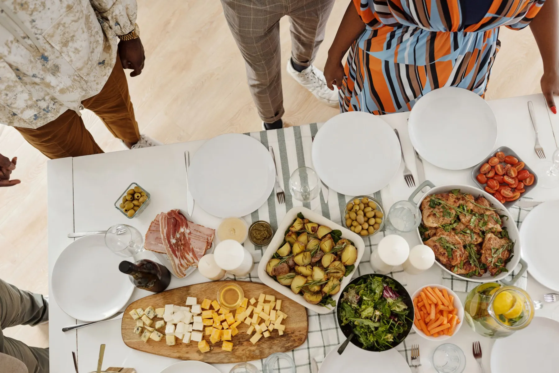 Overhead view of a home table set with roast potatoes, chicken, salad, carrots, tomatoes and cheese as people gather round