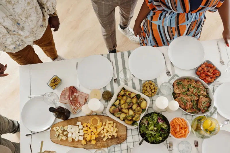 Overhead view of a home table set with roast potatoes, chicken, salad, carrots, tomatoes and cheese as people gather round