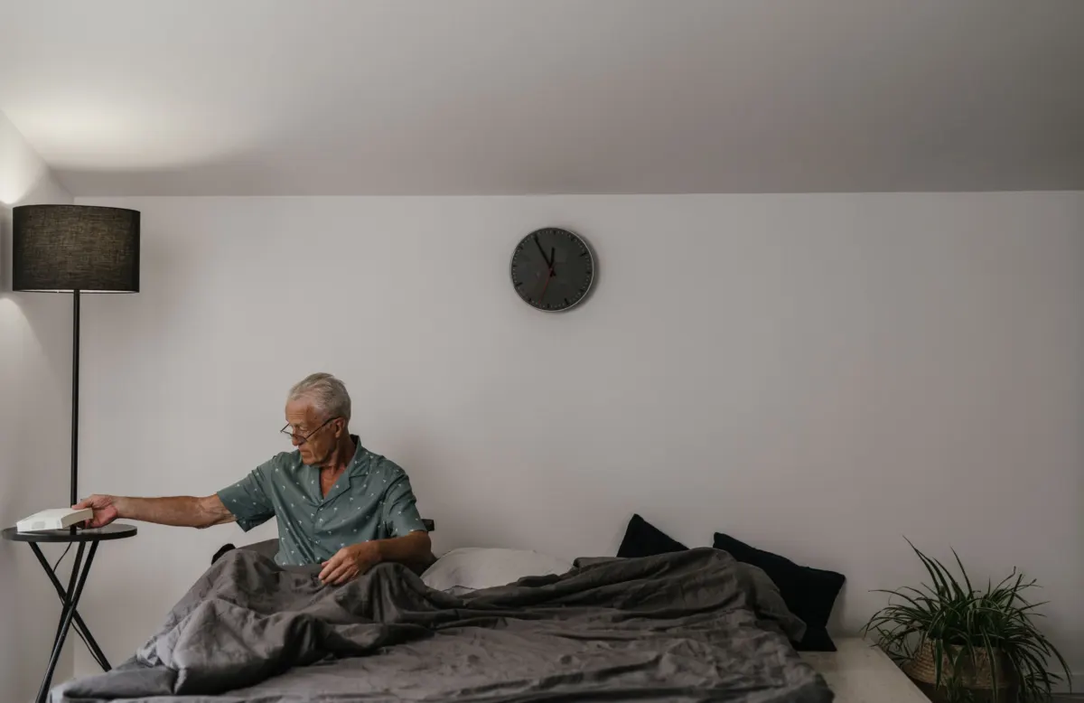 Older man in a short-sleeved shirt sitting up in bed, reaching for a book on a side table beside a floor lamp