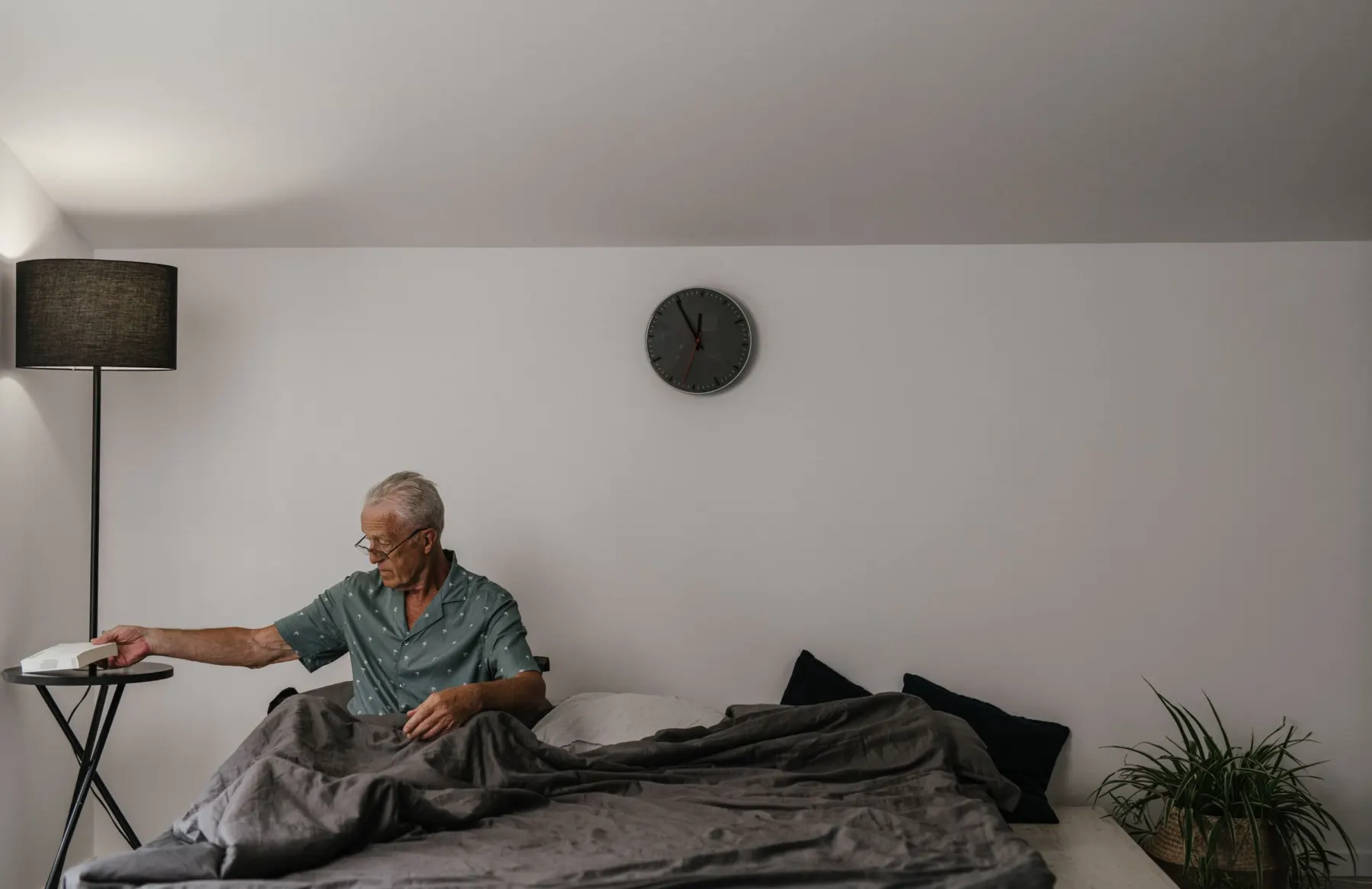 Older man in a short-sleeved shirt sitting up in bed, reaching for a book on a side table beside a floor lamp