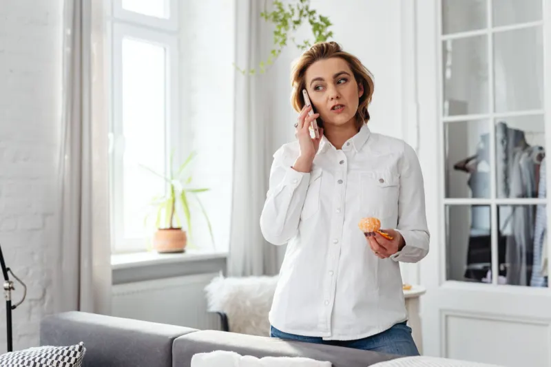 A woman in a white shirt stands in a bright living room talking on the phone, a peeled mandarin in her other hand.