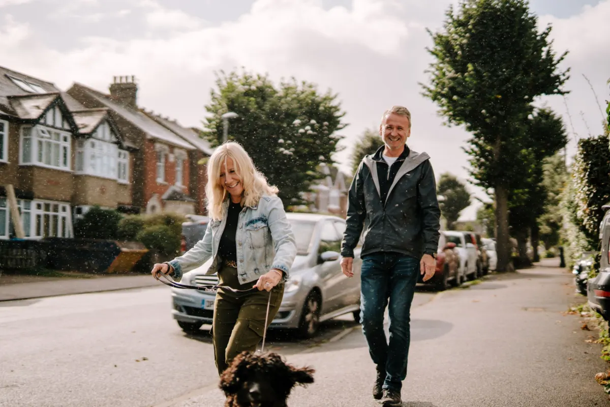 A woman and a man laugh as they walk a dark curly-haired dog along a tree-lined residential street on a bright day.