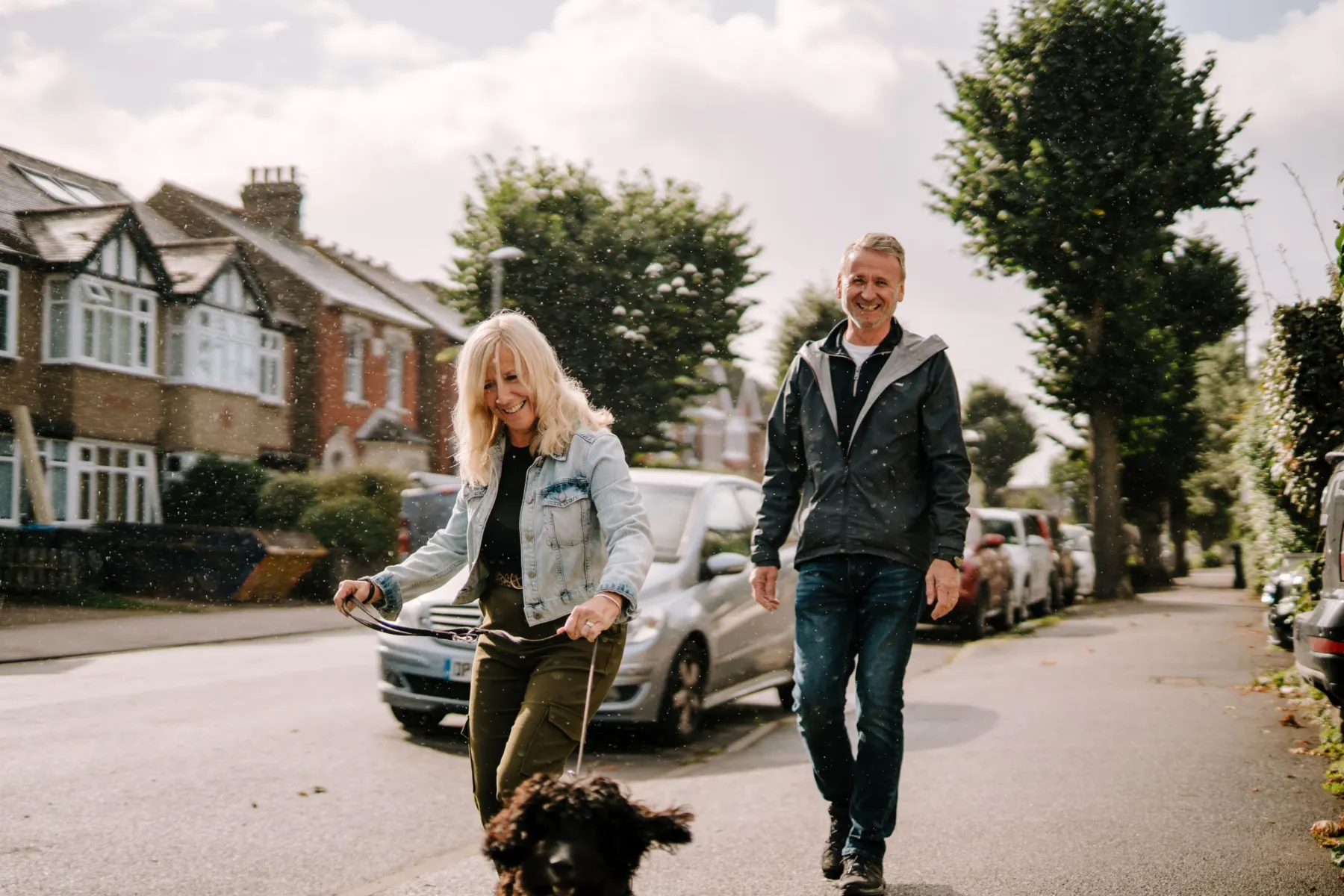 A woman and a man laugh as they walk a dark curly-haired dog along a tree-lined residential street on a bright day.