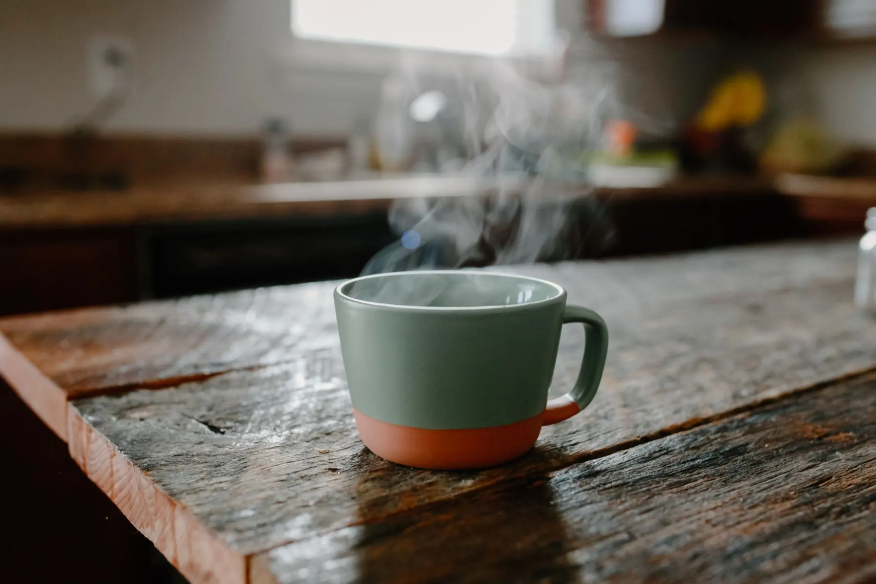 Steaming green and terracotta mug on a worn wooden kitchen worktop, daylight from a window behind
