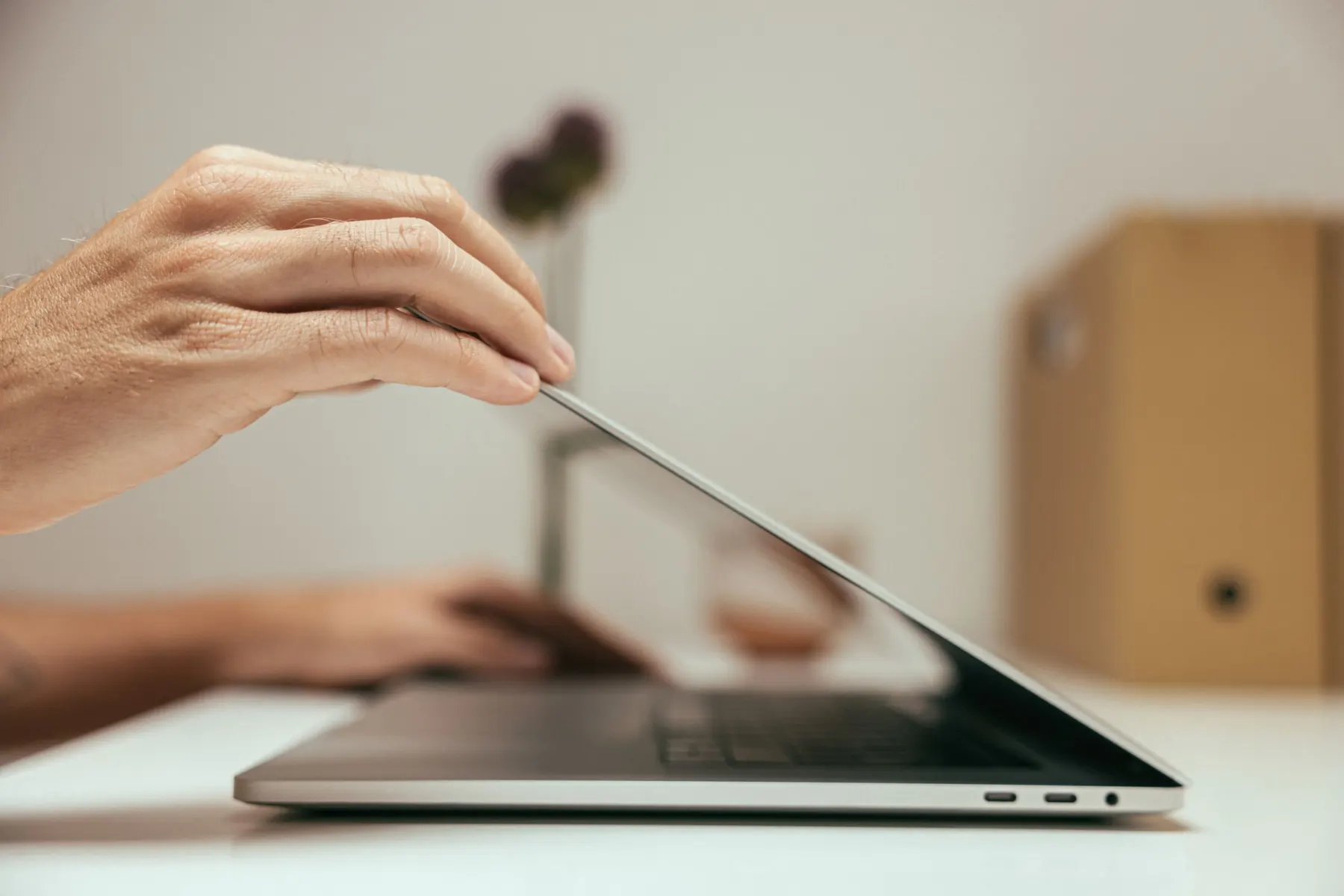 Hand lowering the lid of a laptop on a white desk, a second hand and a cardboard file box blurred behind