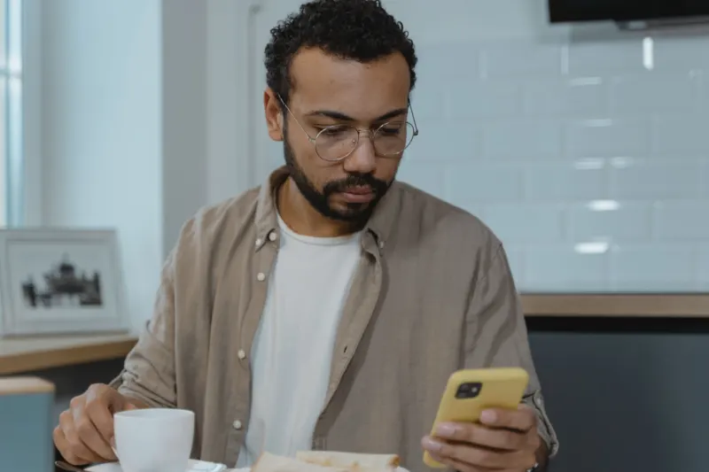 Man with glasses and a beard reading his phone at a kitchen table, a cup and toast in front of him