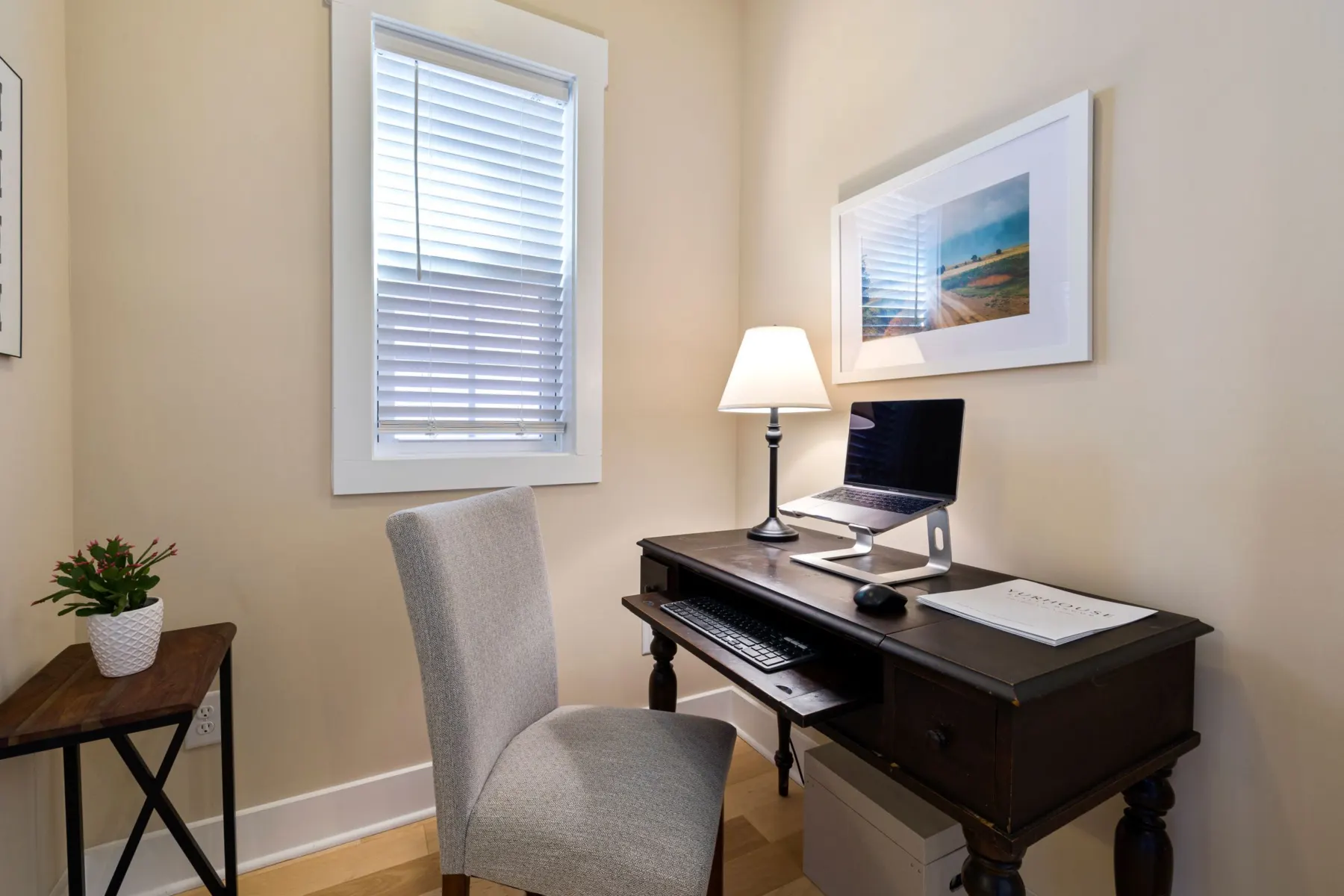 A small home desk by a window: laptop raised on a stand, separate keyboard and mouse, a table lamp and an upholstered chair.