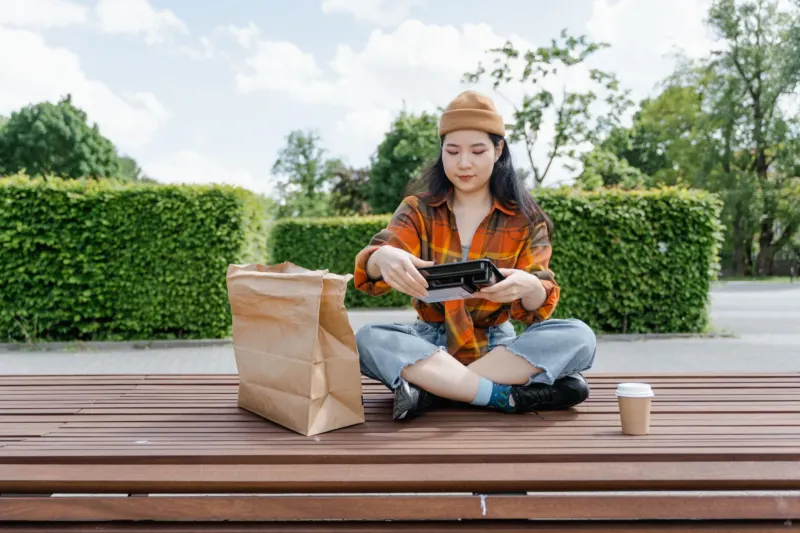 A young woman in a beanie and checked shirt sits cross-legged on a wooden park deck opening a takeaway lunch box beside a paper bag.