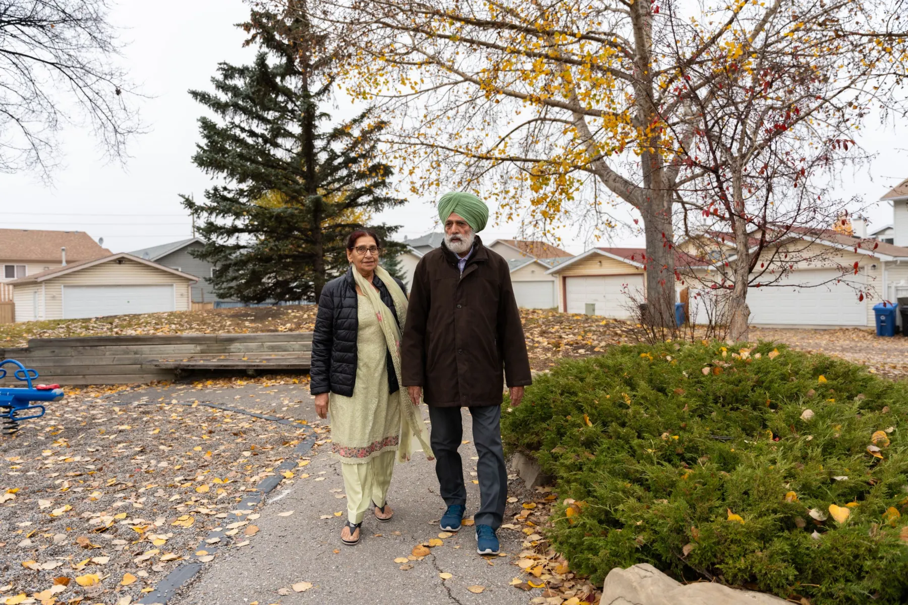 An older couple in everyday coats walk side by side along a leaf-strewn path through a suburban neighbourhood in autumn.