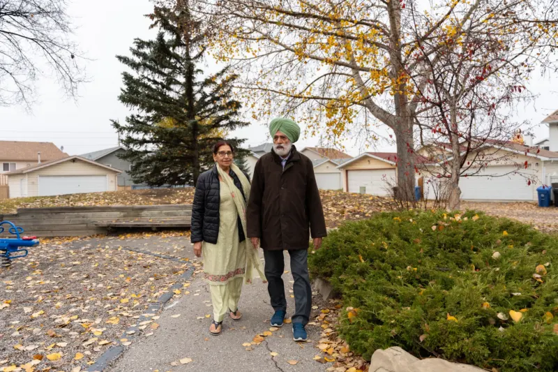 An older couple in everyday coats walk side by side along a leaf-strewn path through a suburban neighbourhood in autumn.