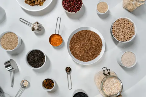 Bowls and scoops of buckwheat, chickpeas, red lentils, kidney beans, brown rice and oats on a white table, seen from above