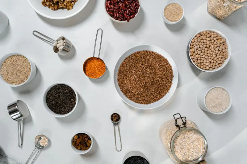 Bowls and scoops of buckwheat, chickpeas, red lentils, kidney beans, brown rice and oats on a white table, seen from above