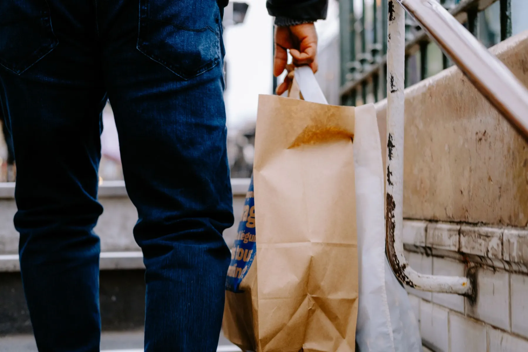 Close view of a person in jeans carrying a full paper shopping bag and a reusable bag up a flight of outdoor steps.
