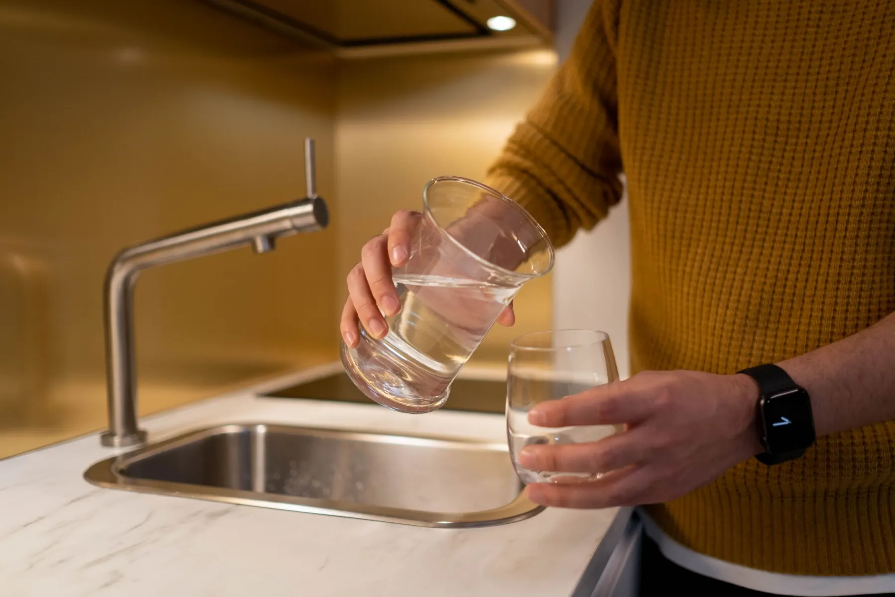 Person in a mustard jumper pouring water from one glass into another beside a kitchen sink