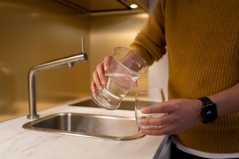 Person in a mustard jumper pouring water from one glass into another beside a kitchen sink