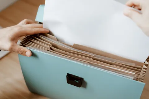 Hands slipping a sheet of paper into a pale blue expanding file with brown card dividers on a wooden table