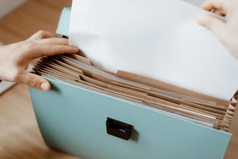 Hands slipping a sheet of paper into a pale blue expanding file with brown card dividers on a wooden table