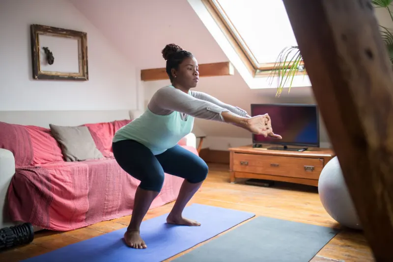 A woman does a squat on a mat between the sofa and the television in a small attic living room lit by a skylight.