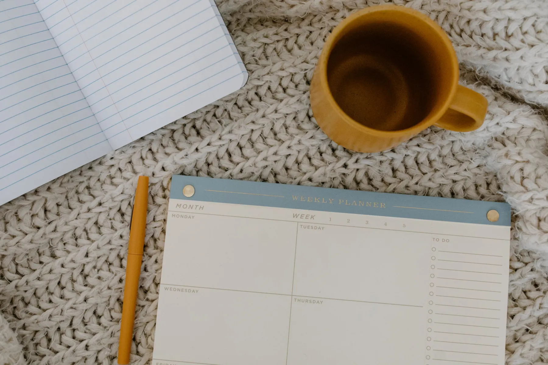 A blank weekly planner pad, a mustard pen, an empty mustard mug and an open lined notebook lie on a chunky cream knitted blanket.