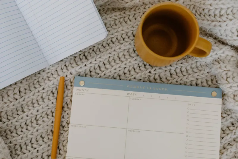 A blank weekly planner pad, a mustard pen, an empty mustard mug and an open lined notebook lie on a chunky cream knitted blanket.