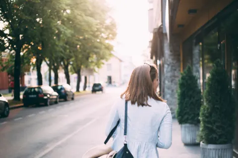 A woman seen from behind walks along a tree-lined street in low morning sun, a bag over her shoulder and a coat on her arm.