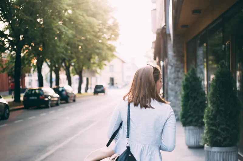 A woman seen from behind walks along a tree-lined street in low morning sun, a bag over her shoulder and a coat on her arm.