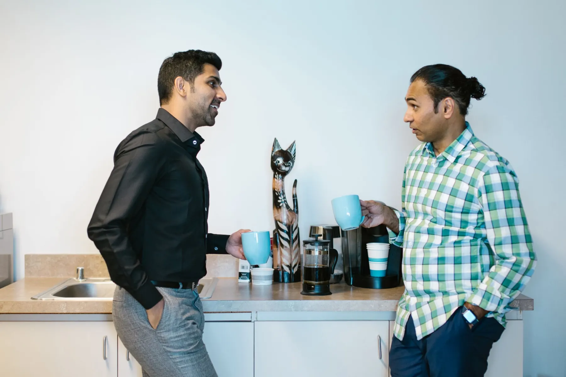 Two men lean against an office kitchen counter holding blue mugs and chatting beside a cafetière and a wooden cat figure.