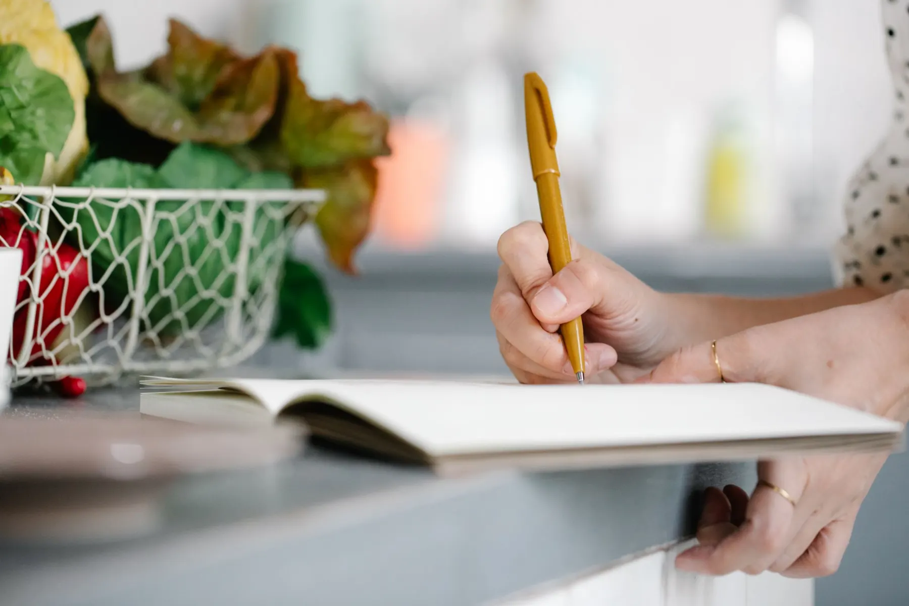 Hand writing in an open notebook on a kitchen counter beside a wire basket of lettuce and vegetables