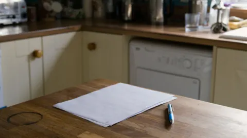 Blank sheets of paper and a blue pen on a worn wooden kitchen table, cream cupboards behind