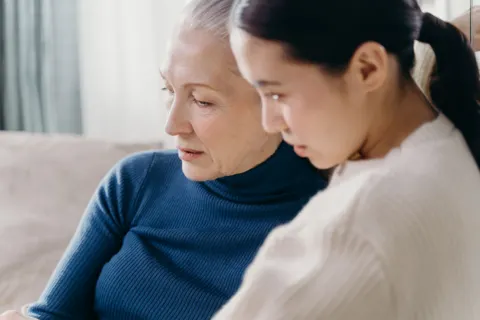An older woman in a blue jumper and a younger woman in a cream jumper sit close on a sofa, both looking down