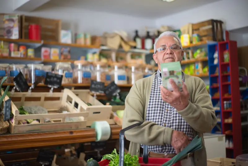 Older man with glasses in a small grocery shop holding up a carton to read it, a basket of vegetables on his arm