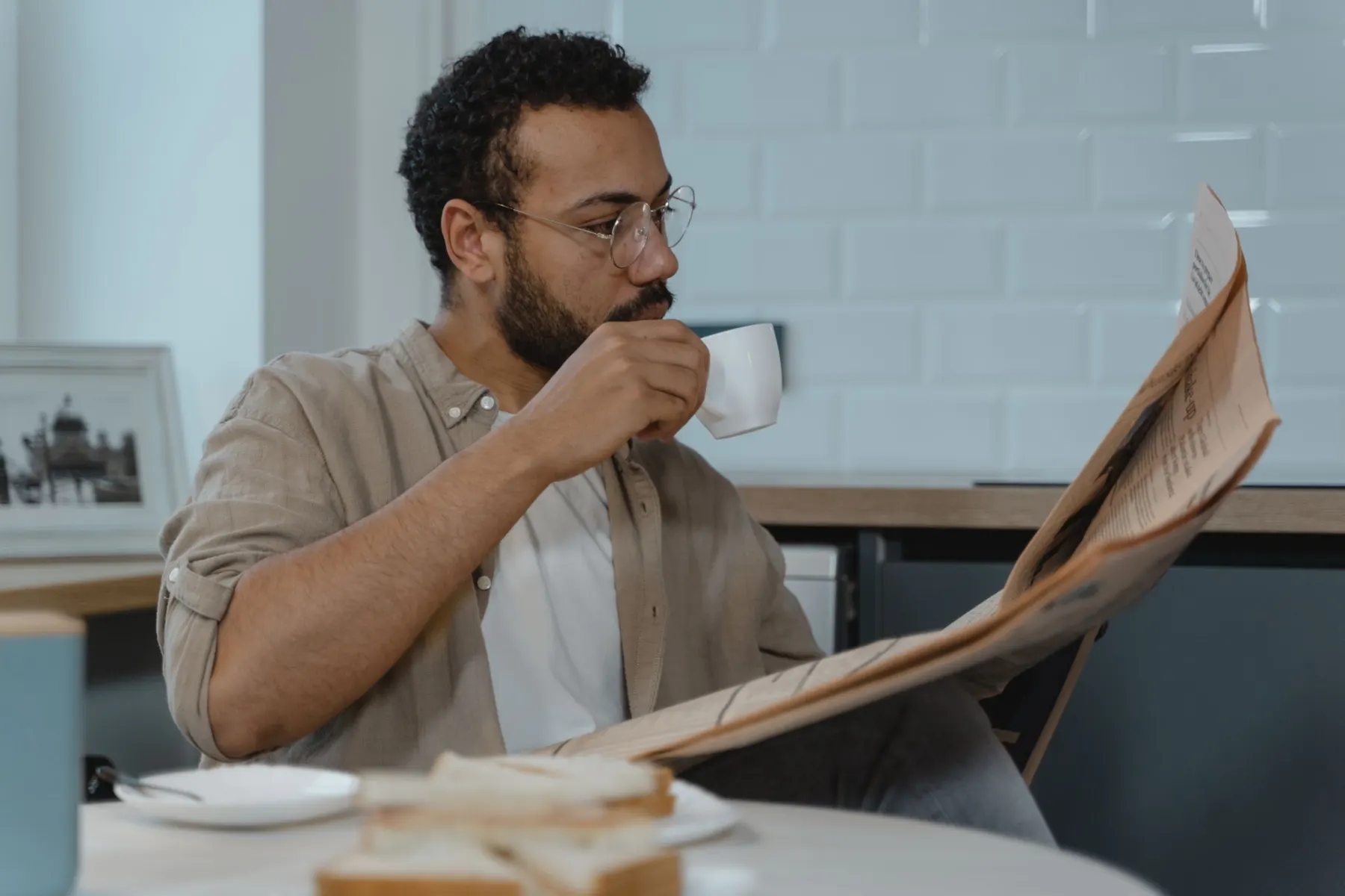 A bearded man in round glasses sips from a white cup while reading a newspaper at a kitchen table