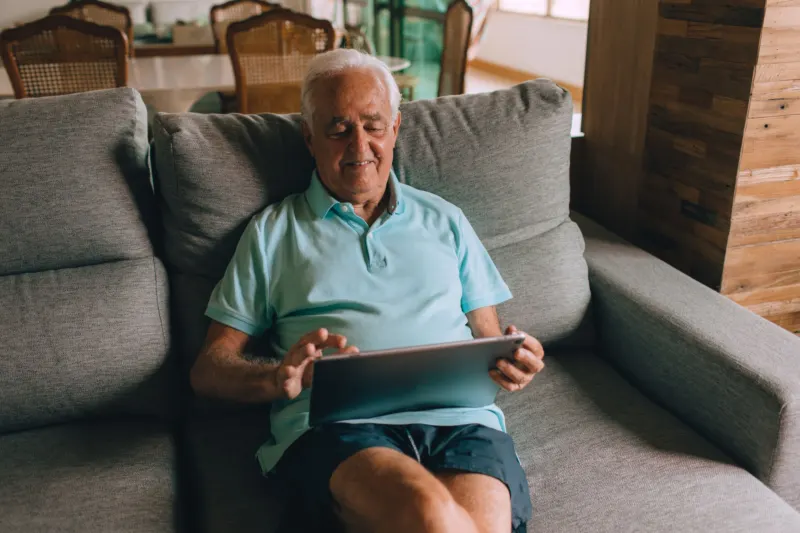 An older man with white hair sits back on a grey sofa at home, tapping a tablet resting on his lap