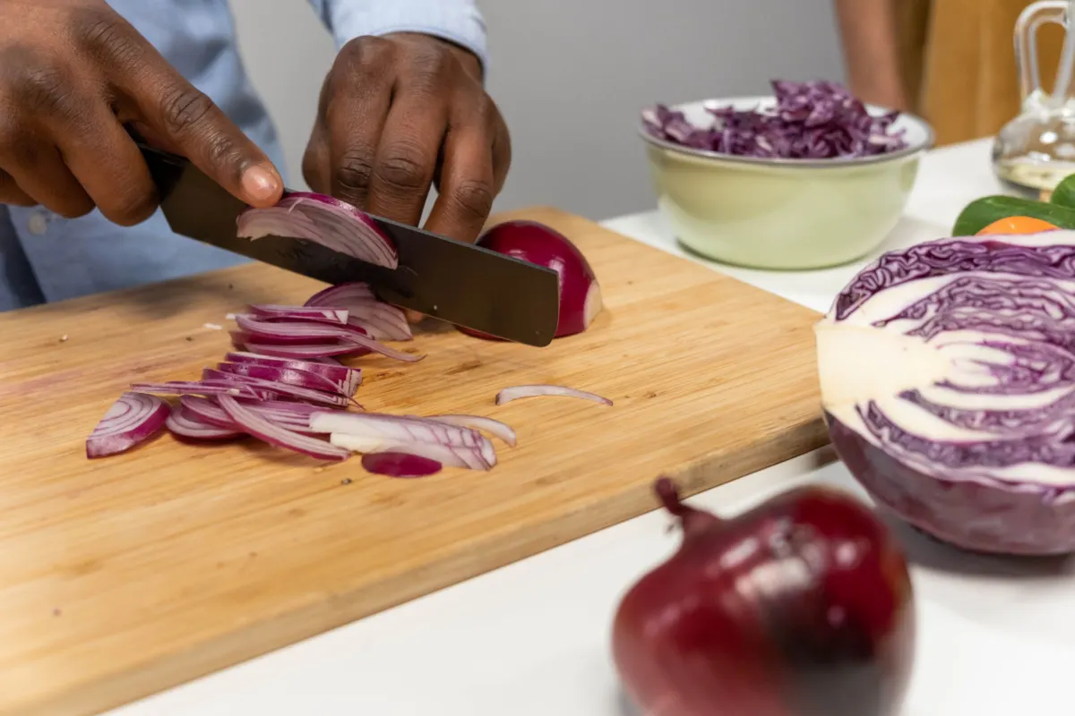 Hands slicing a red onion on a wooden board, with a bowl of shredded red cabbage and half a cabbage alongside