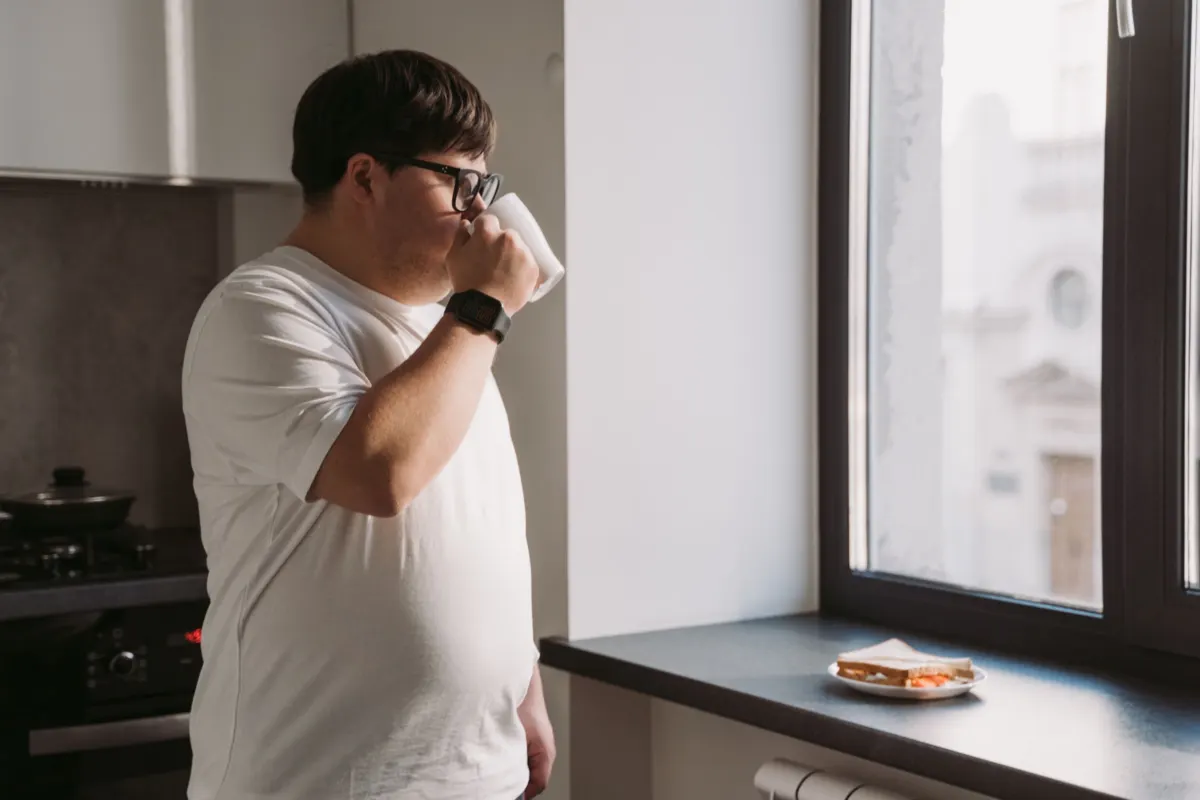 A man in glasses and a white T-shirt drinks from a white mug in his kitchen, facing a bright window with a sandwich on the sill.