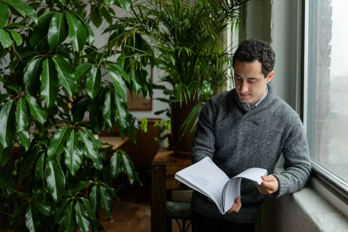 A man in a grey jumper stands by a window leafing through a printed booklet, large green houseplants behind him
