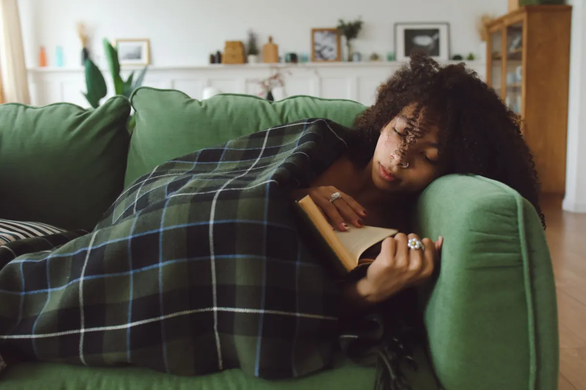 Woman with curly hair dozing on a green sofa under a tartan blanket, an open book resting under her hand