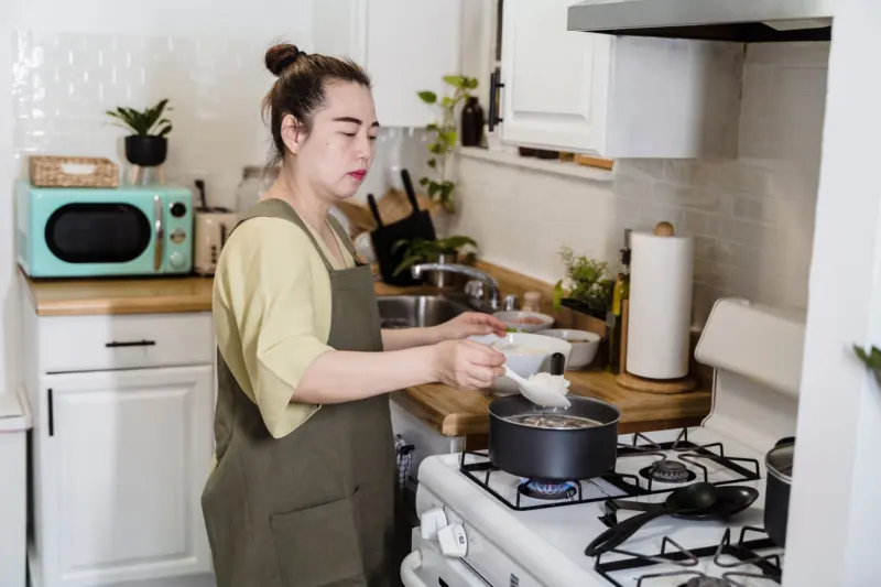 Woman in an apron spooning rice into a pot on a gas hob in a small home kitchen