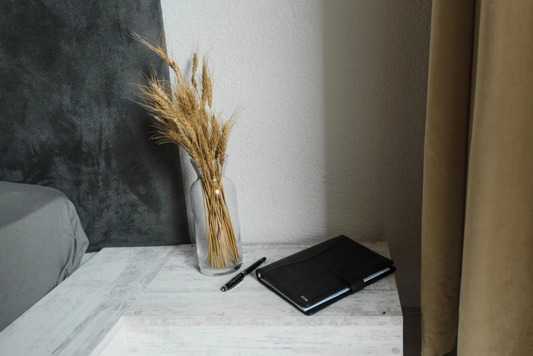 Closed black planner and a pen on a pale bedside table next to a glass vase of dried wheat, bed edge at left