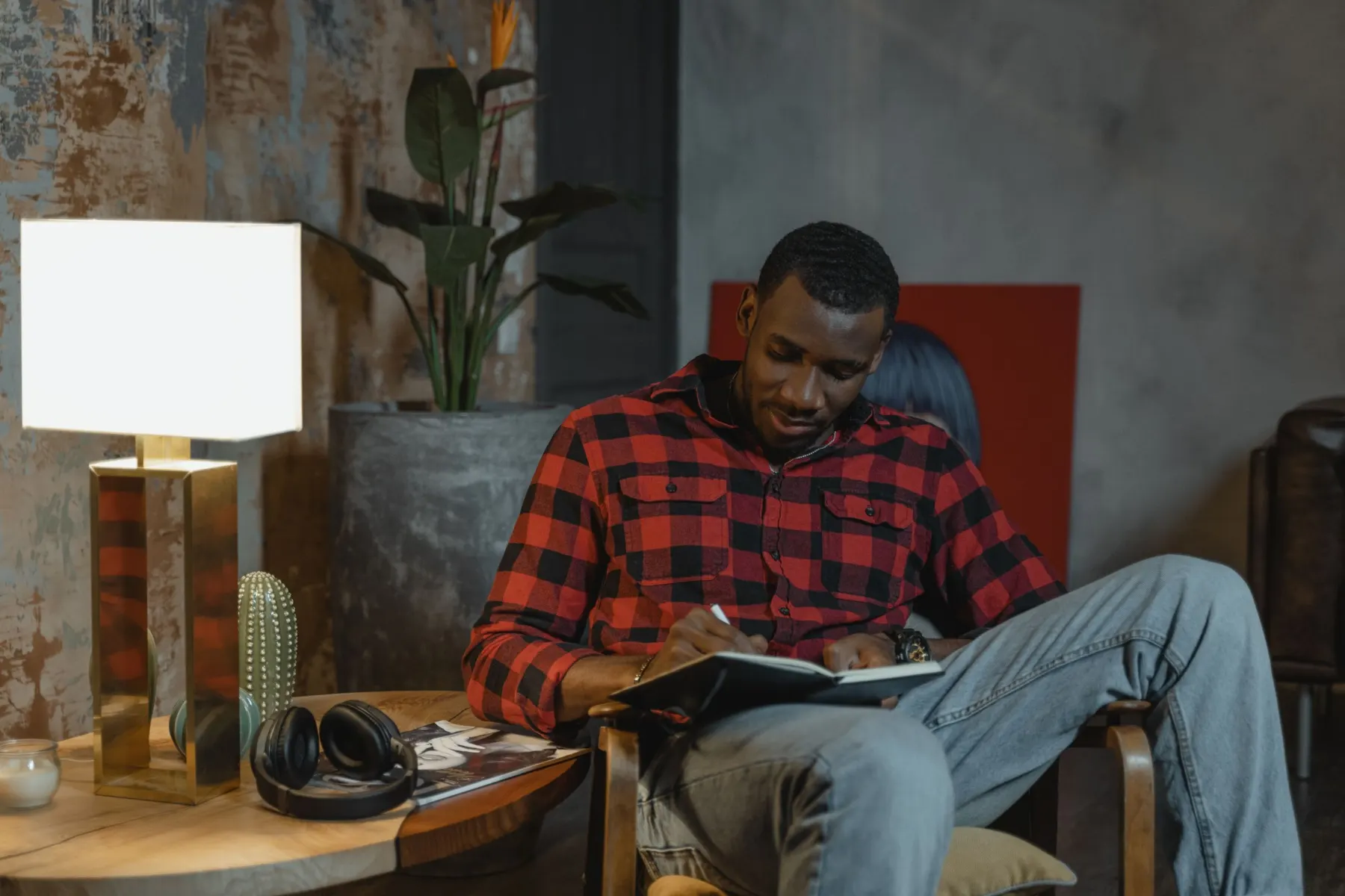 A man in a red checked shirt sits in an armchair beside a lit table lamp, writing in a notebook resting on his knee.
