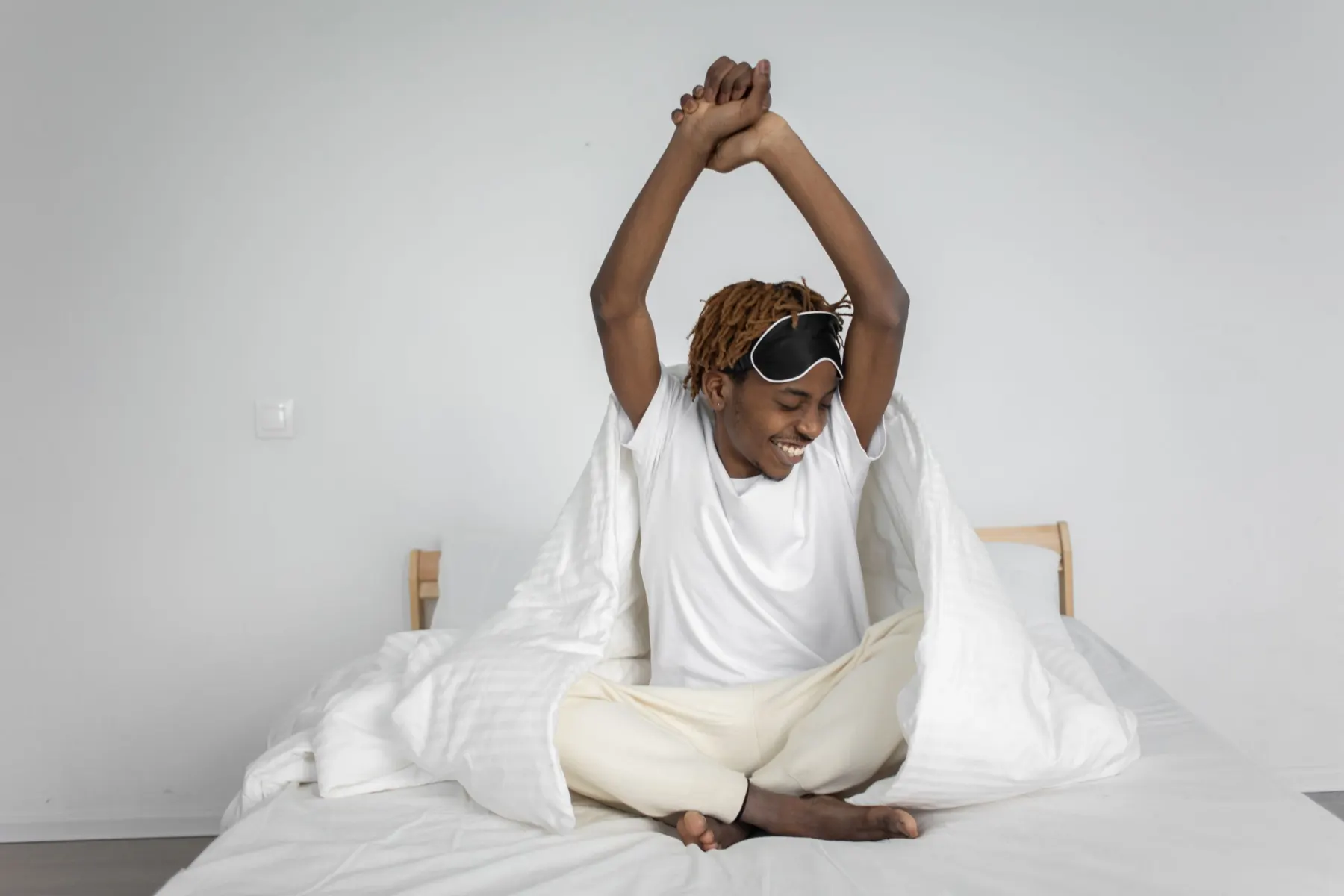 Young man sitting cross-legged on a bed in a white T-shirt, smiling and stretching his arms overhead, sleep mask pushed up