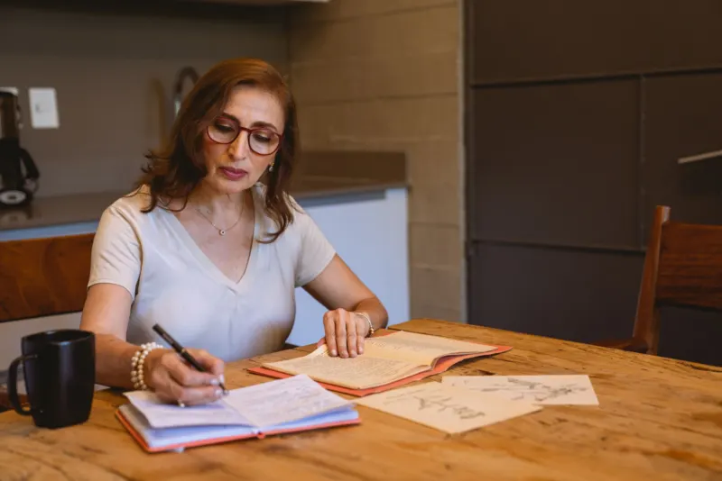 Woman in glasses writing in a notebook at a wooden kitchen table, an open booklet and a dark mug beside her