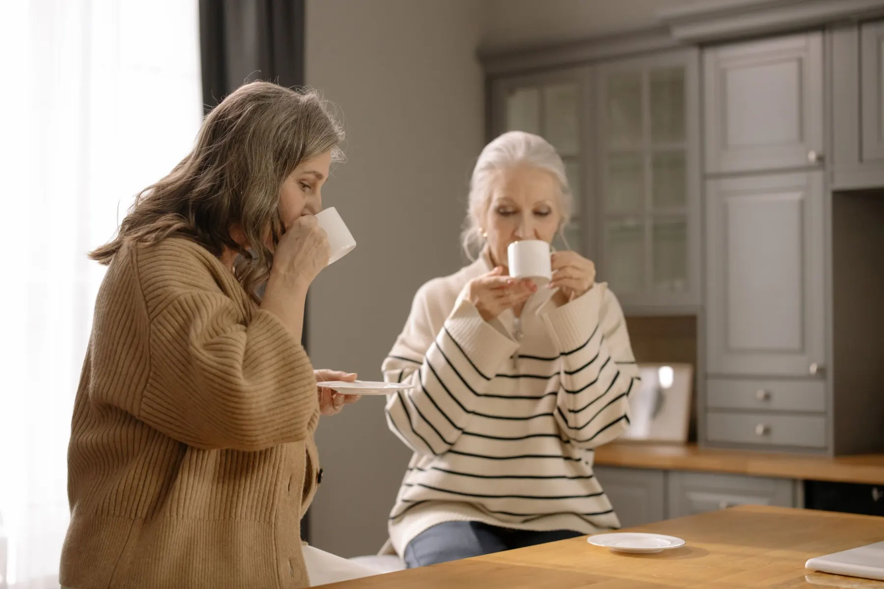 Two older women sit together at a wooden kitchen counter, each sipping from a white cup in soft window light.