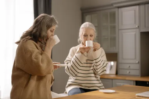 Two older women sit together at a wooden kitchen counter, each sipping from a white cup in soft window light.
