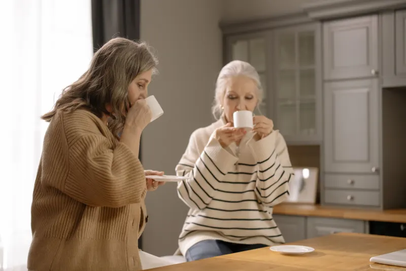 Two older women sit together at a wooden kitchen counter, each sipping from a white cup in soft window light.